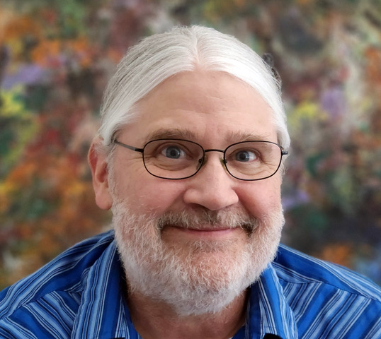 A man with white pulled back hair and beard wearing wire glasses and a blue striped shirt smiling brightly.