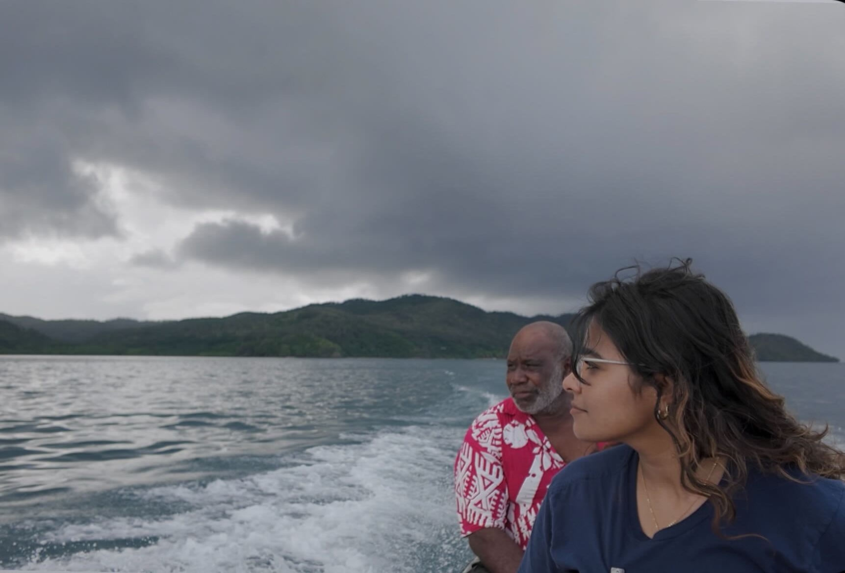A woman with dark hair pulled back wearing glasses on a boat with a man in profile with islands in the backgroud.