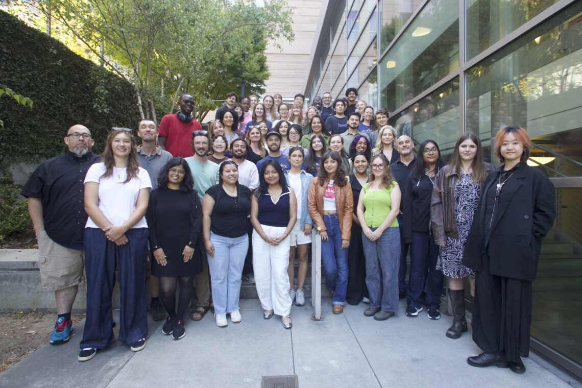 A group of journalism students assembled on the steps of a building with a reflection of them on the pane to their left side.