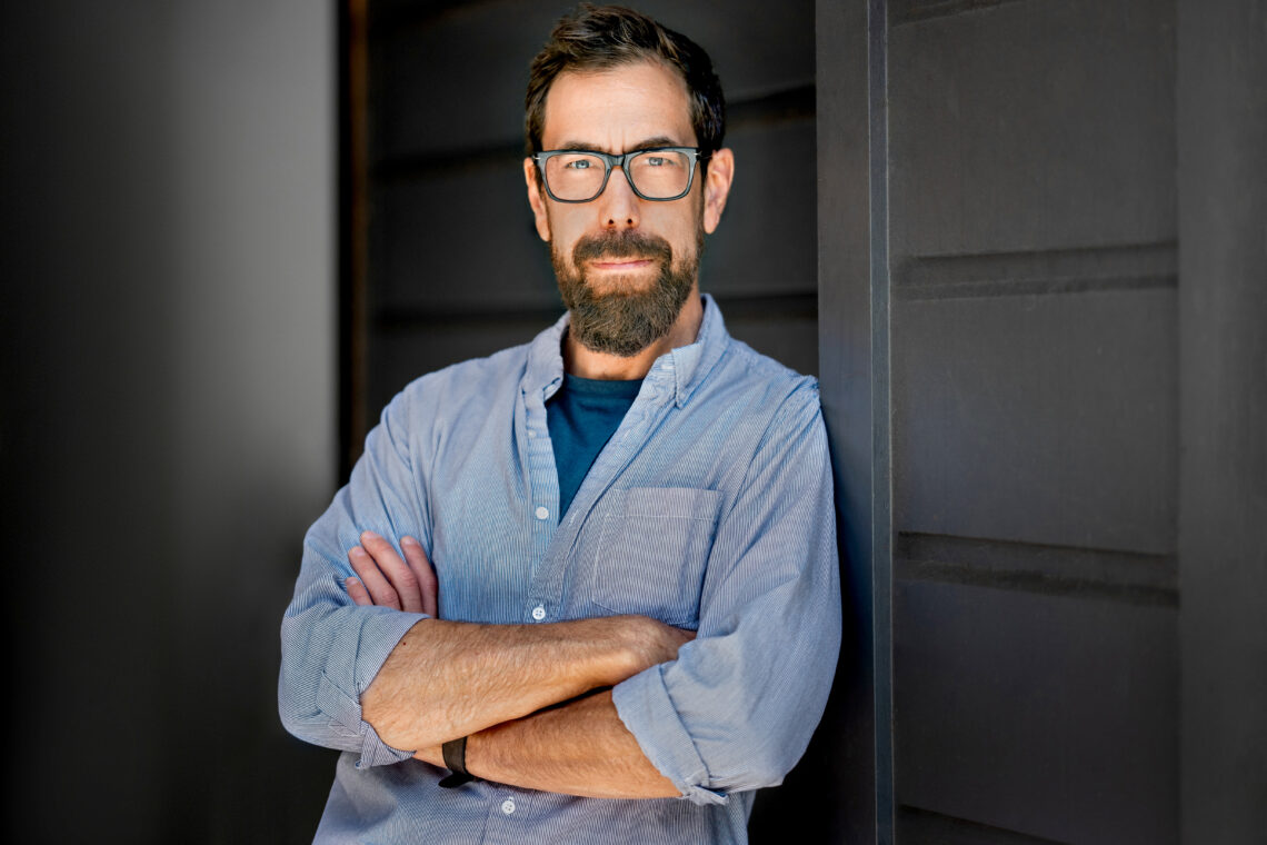 A man with brown hair and beard wears glasses and a blue botton down with his arms crossed.