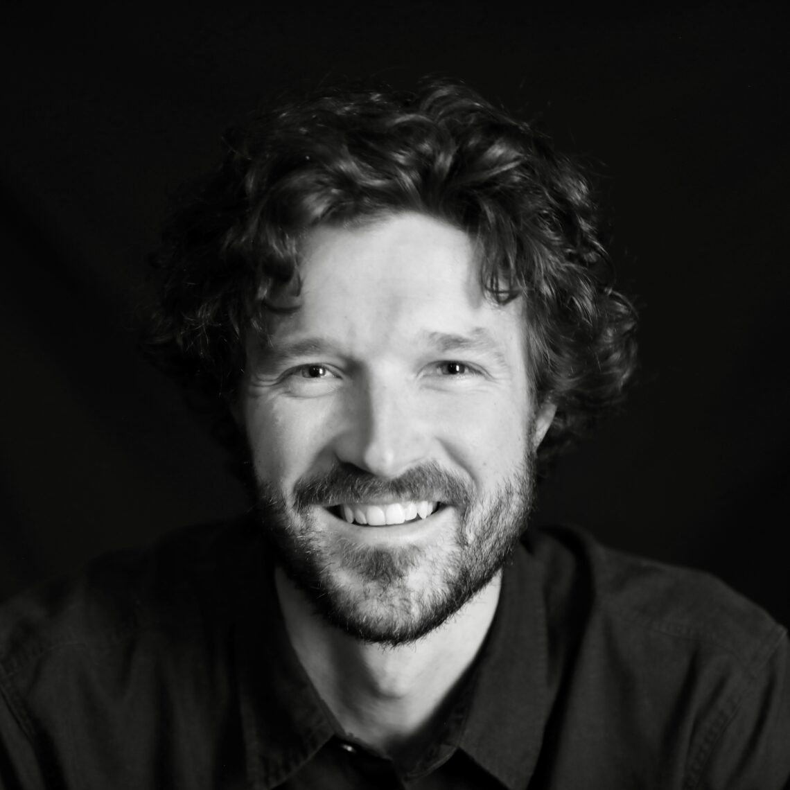 Black and white headshot of a young male filmmaker with brown wavy hair and a mustache and beard smiling.