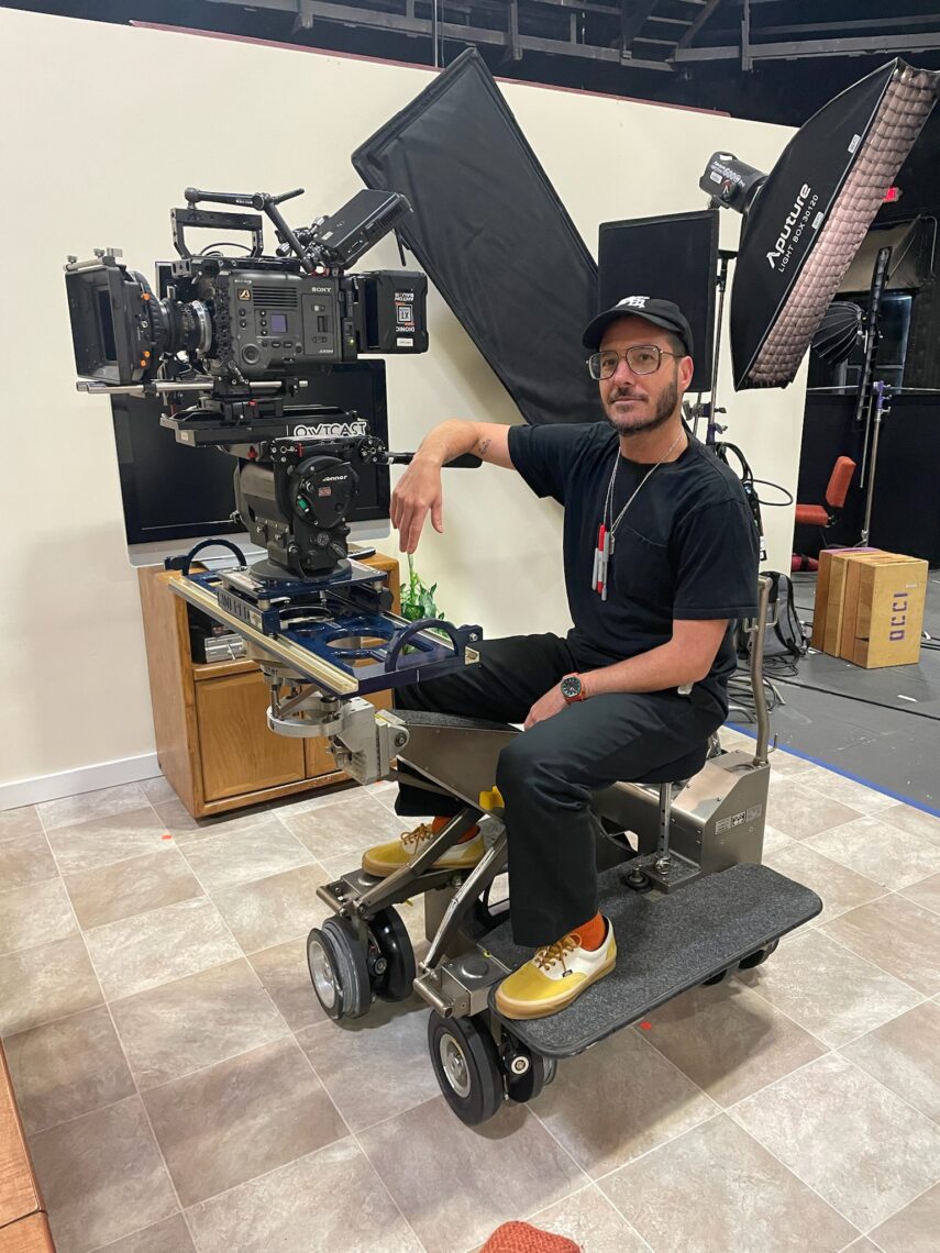 A man wears a black baseball hat, t-shirt and jeans while sitting on a sophisticated movie camera set-up.