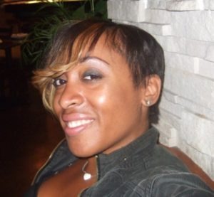 A woman with short hair, featuring blond highlights on one side, smiling at the camera. She is wearing a dark jacket and a necklace with a heart-shaped pendant. The background shows a stone wall and some greenery. Carah Herring's serene expression radiates warmth.