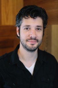 A man with wavy dark hair and a beard, presumed to be David Cohn, is seen from the chest up, wearing a black collared shirt over a white t-shirt. He is standing in front of a background with wooden panels and has a neutral expression.