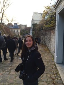 Alison Vayne, with long hair and wearing a dark jacket, stands on a cobblestone path, smiling at the camera. She has a camera slung around her neck. Other people are walking ahead on the path, which is lined with stone walls and bare trees.