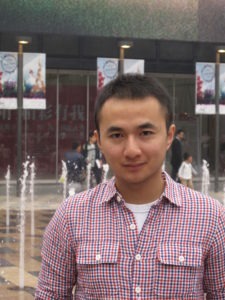 A young man in a red and blue checkered shirt stands in front of a water fountain with a backdrop of a modern building. There are several banners hanging on poles behind him. Leo Zou looks directly at the camera with a slight smile.