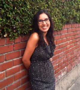 A woman with long dark hair and glasses is smiling while leaning against a brick wall covered with green ivy. Guisel Contreras is wearing a sleeveless black and white polka dot dress and has her hands behind her back.