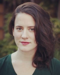 Katherine Rose, a woman with wavy, dark brown hair and light skin, is looking steadily at the camera. She is wearing a dark green top and has a calm expression on her face. The background features blurred greenery, highlighting her serene presence.