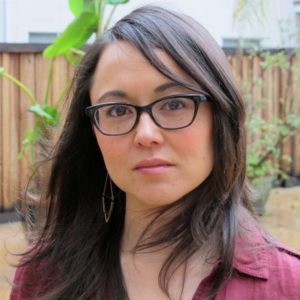 Jun Stinson, a woman with long brown hair and glasses, stands in an outdoor setting with a wooden fence and greenery in the background. Wearing a maroon shirt, she has a serious expression on her face as a slight breeze moves her hair.