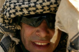 Close-up photograph of Ethan Baron wearing a patterned headscarf and dark sunglasses. The individual has a smile and is framed by the fabric of the headscarf surrounding their face.