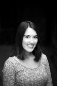 A black and white portrait of Noelia Gonzalez, a young woman with shoulder-length dark hair. She's smiling and wearing a short-sleeved, textured top. The background is dark and out of focus, drawing attention to her face.