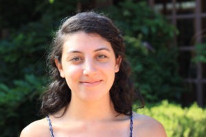 A young woman with curly dark hair, Katherine Harloe, smiles softly at the camera. She is wearing a sleeveless top and stands in front of a leafy green background, suggesting an outdoor setting on a sunny day.