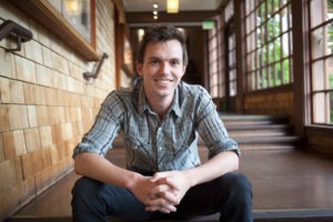 James Reddick, a man with short dark hair, wearing a gray plaid shirt and black pants, sits on a step in a well-lit hallway. He is smiling and has his hands clasped in front of him. The hallway has wooden walls and large windows along one side.