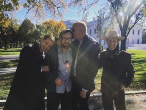 Four men are standing together outside on a sunny day with trees and a white building in the background. In this joyful alumni portrait, one man, identified as Jeffrey Plunkett, is being hugged and kissed on the head by two others, while a producer wearing a hat stands smiling to the right. Everyone appears cheerful.
