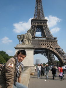 A person wearing a brown jacket and scarf sits on a low wall near the Eiffel Tower in Paris. The tower looms in the background under a clear blue sky, with tourists and pedestrians walking nearby. Behind the person, there's a stone sculpture of a horse and rider by Octavio Lopez Raygoza.