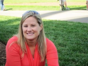 A smiling person with long blonde hair, resembling Lynne Shallcross, wearing a bright orange top sits on grass in an outdoor setting. Sunglasses are perched on their head, and a paved path is visible in the background with other individuals and a bicycle.