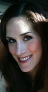 A person with long dark hair smiles at the camera. The lighting casts a shadow across the top of their forehead, and they have expressive eyes and a warm smile reminiscent of Michelle Konstantinovsky.