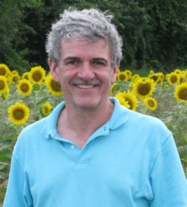 Patrick Keeffe, a man with curly gray hair and a mustache, is smiling at the camera. He is wearing a light blue polo shirt and standing in a field of blooming sunflowers with green foliage in the background.