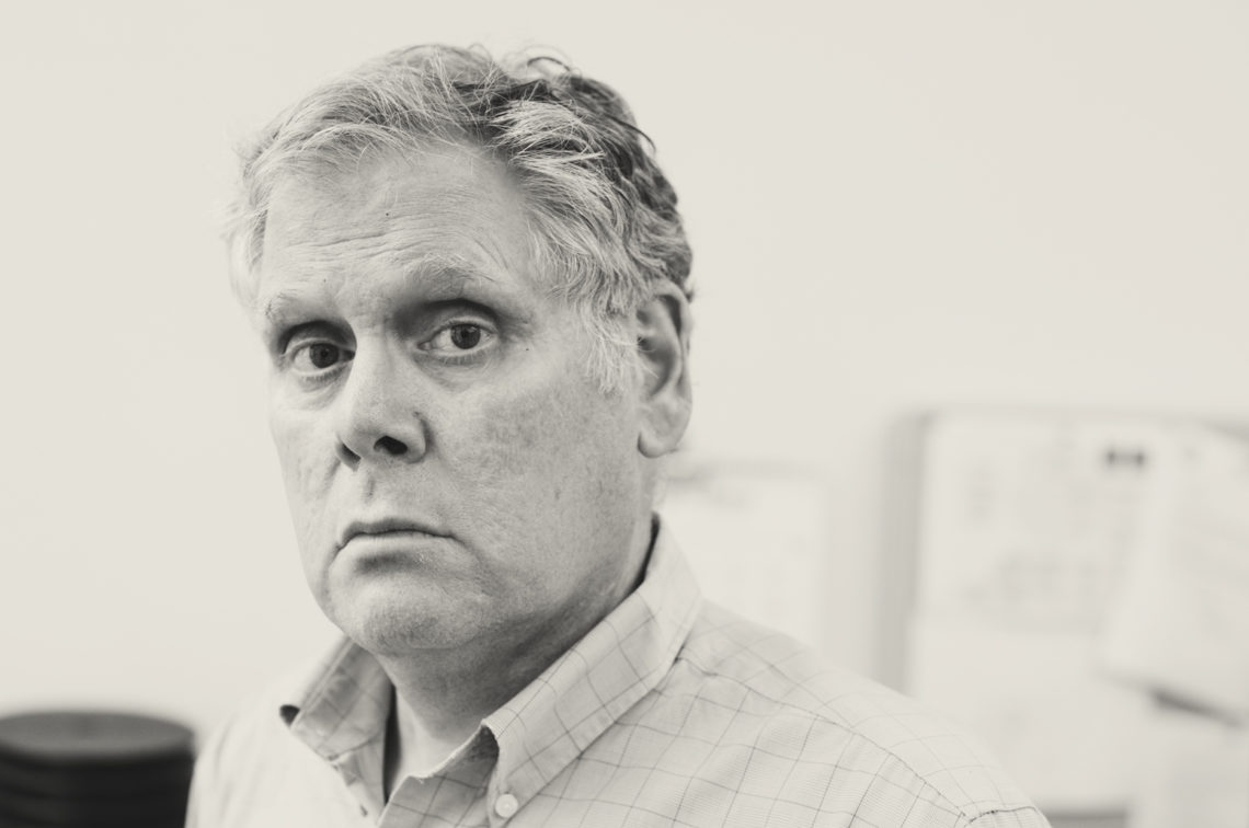A grayscale lecturer portrait of a man with short, graying hair, wearing a checkered shirt. Thomas Peele is looking directly at the camera with a neutral or serious expression. The background is out of focus and appears to be an office setting with papers or bulletin boards visible.
