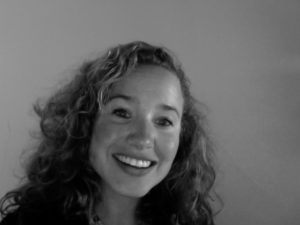 Black and white photo of a smiling person with medium-length curly hair. They are looking directly at the camera. The background is plain and unadorned, perfectly highlighting Caroline Bins' natural charm.