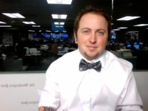 A person with short hair and earrings is seated in a brightly lit office, wearing a white shirt and a black bow tie with a pattern. The office, reminiscent of James Buck's workspace, has multiple computer monitors in the background. The person is looking directly at the camera, smiling slightly.