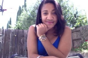 A woman with long, dark hair is smiling and leaning on her arm. She is wearing a blue sleeveless top and a gold wristwatch. In the background, there is a wooden fence, green foliage, and a basketball hoop. The scene appears to be outdoors on a sunny day, perhaps capturing Trenise Ferreira in her element.