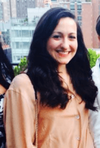 Emily Thomas, a woman with long dark hair, is smiling while wearing a beige top. She stands outdoors with buildings and greenery visible in the background.
