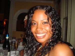 A woman with long curly hair and a big smile, who closely resembles Shikiri Hightower, sits at a table in a restaurant. She is wearing a strapless top and a necklace. There are drinks and wine bottles on the table, and other diners are visible in the background.