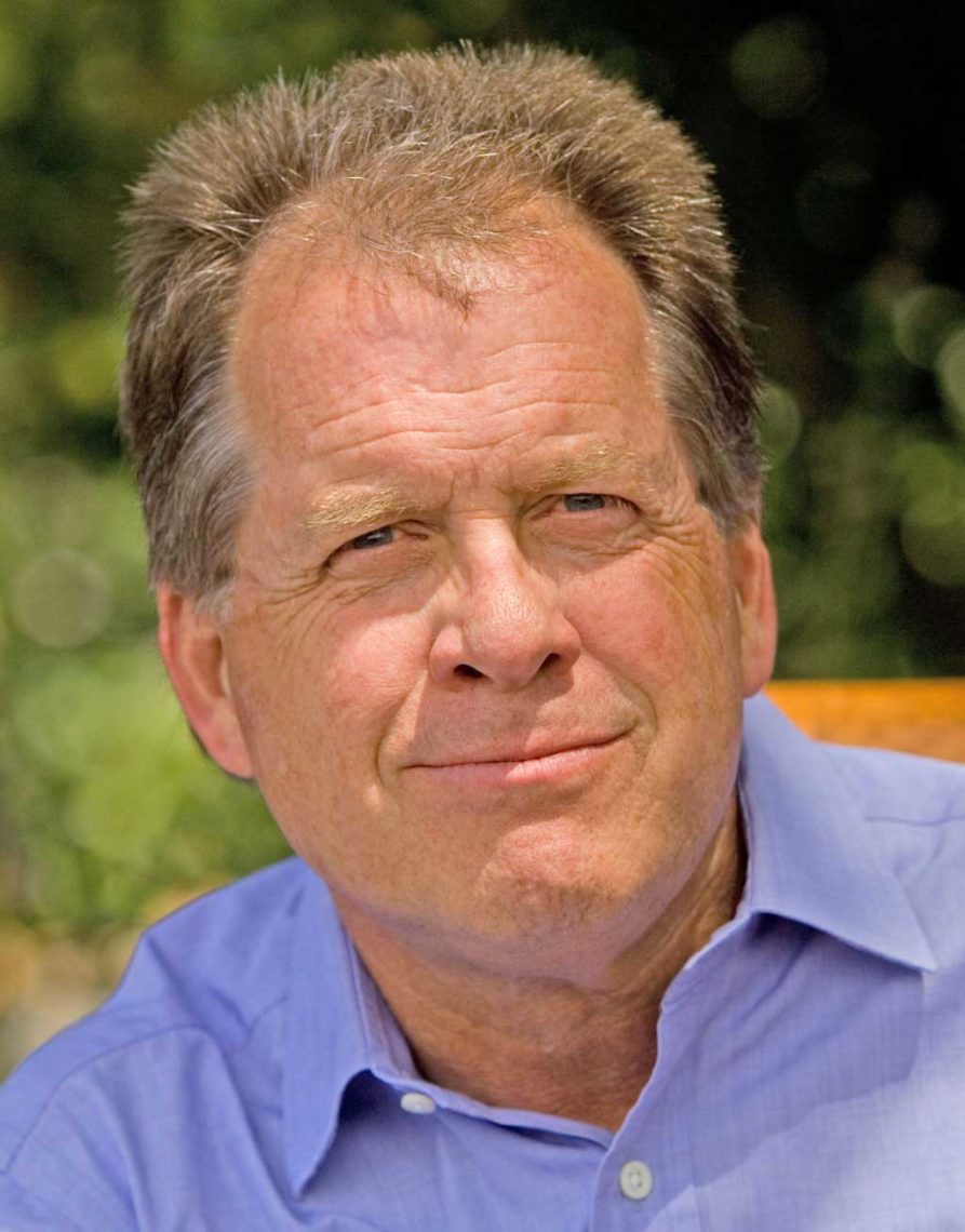 A middle-aged man with short, graying hair is pictured outdoors. He is wearing a light blue collared shirt and is looking directly at the camera with a slight smile, reminiscent of Timothy Ferris. The background is blurred, showing greenery.