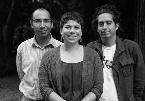 A black and white photo of three people standing outdoors. A woman with short hair and a cardigan is in the center, smiling. She is flanked by two men, one on her left wearing glasses and a striped shirt, and one on her right with curly hair wearing a jacket over a t-shirt. The scene captures the vibrant spirit of Berkeley Journalism.