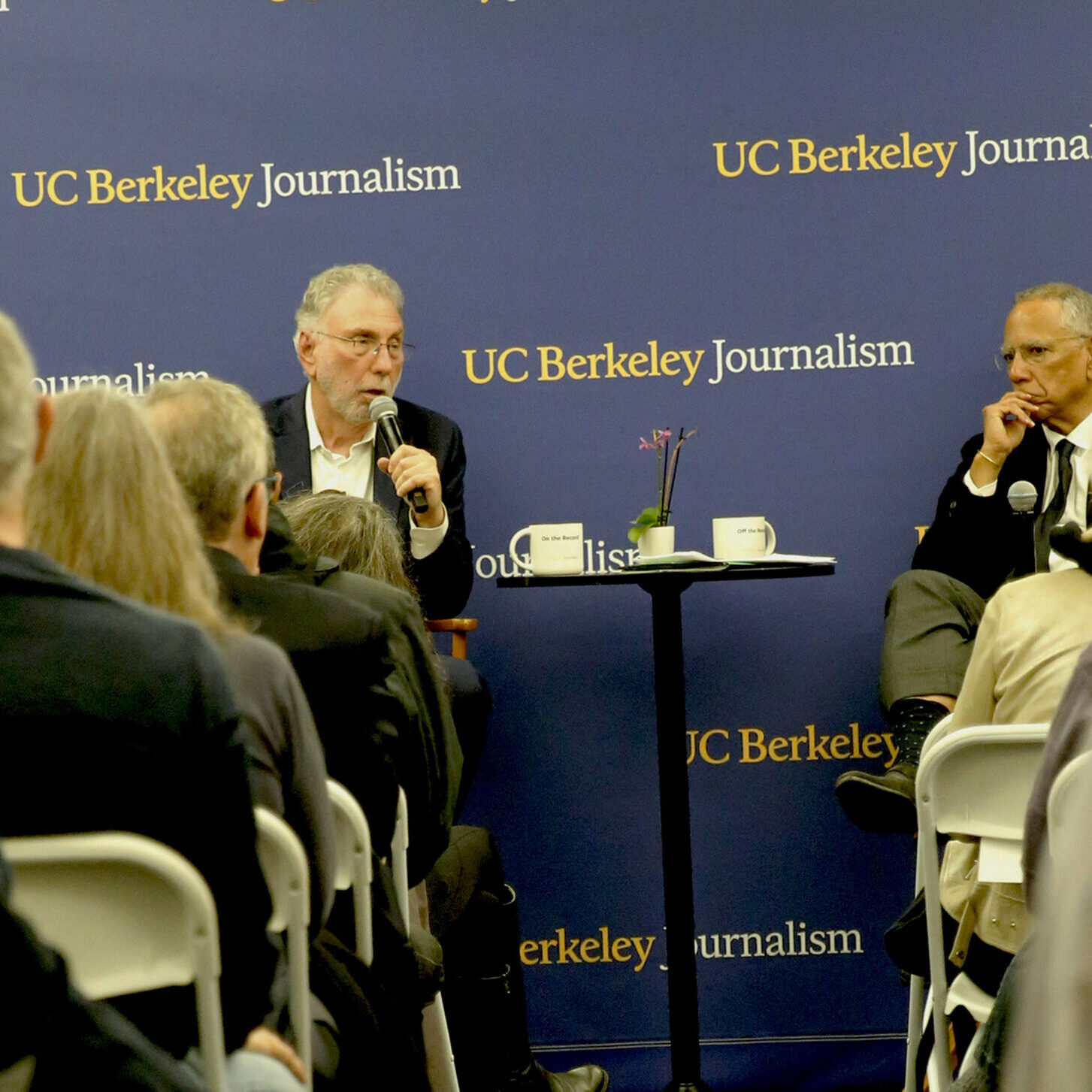 Marty Baron and Dean Baquet talking at North Gate Hall. The photo is taken from the back of the room down the aisle so that you see people on both sides. The backdrop is blue with the words UC Berkeley Journalism in a step and repeat pattern.