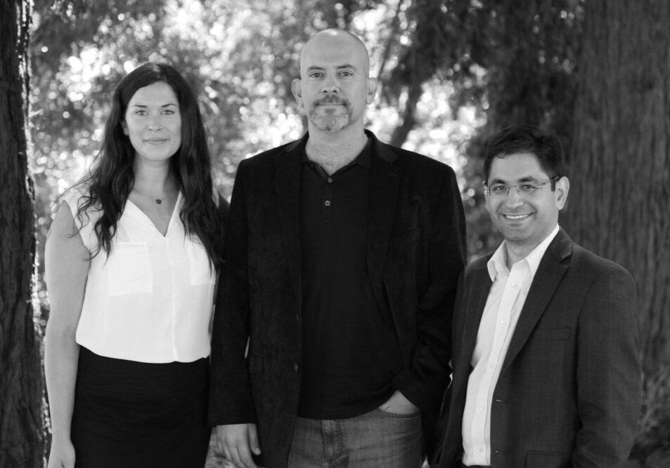 Three individuals stand outdoors, positioned in front of trees. From left to right, a woman with long dark hair wearing a white blouse and black skirt, a man with a shaved head and beard in a dark blazer, and a man with glasses in a suit smiling. Their camaraderie radiates the spirit of Berkeley Journalism.