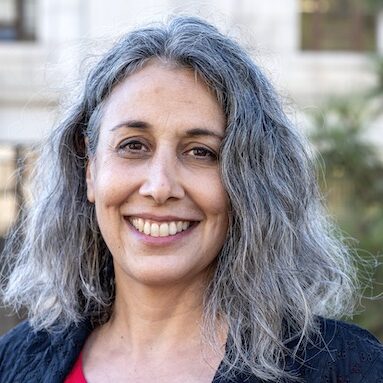 A woman with long, wavy, gray hair smiles at the camera. She is wearing a red top and a black jacket. Against a backdrop of blurred greenery and a building, she exudes confidence—a true embodiment of Berkeley Journalism.