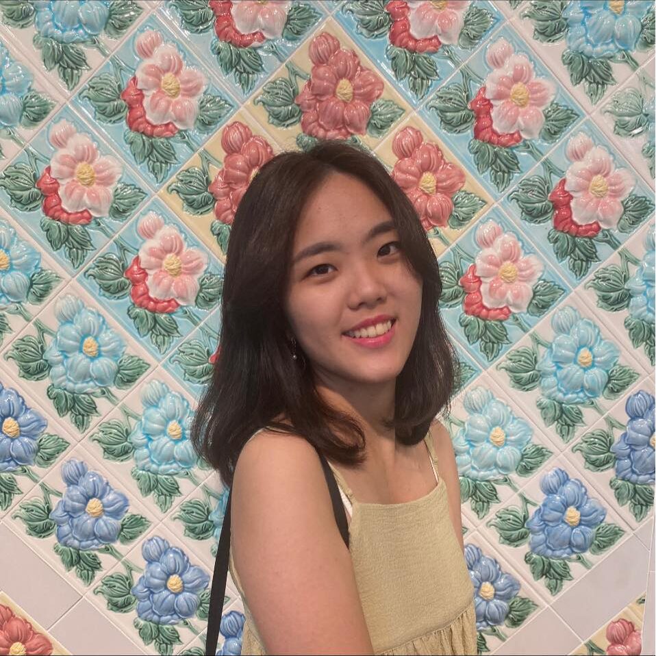 Hailey-Wang-1 A woman with shoulder-length hair smiles in front of a floral tile wall featuring colorful red, pink, blue, and green flowers.