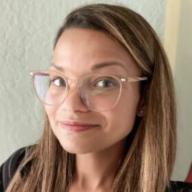 A woman with long straight hair and glasses is smiling at the camera. She wears a light-colored top and stands indoors, against a light-colored wall, exuding warmth that's perfect for welcoming new admissions.