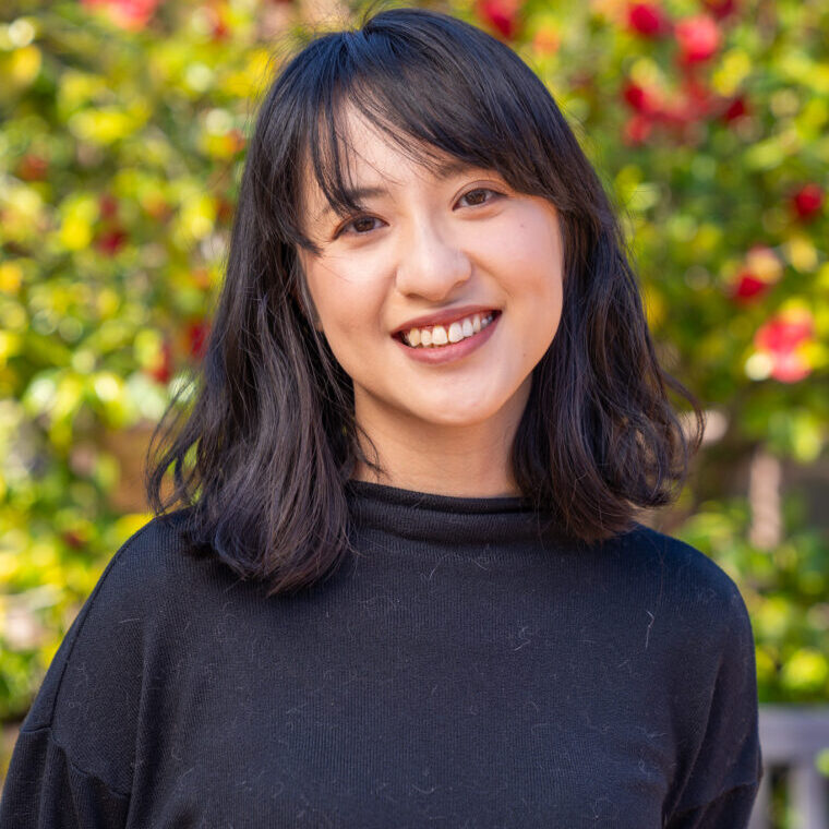 Woman smiling has dark hair and a black sweater with a flowering tree behind her.