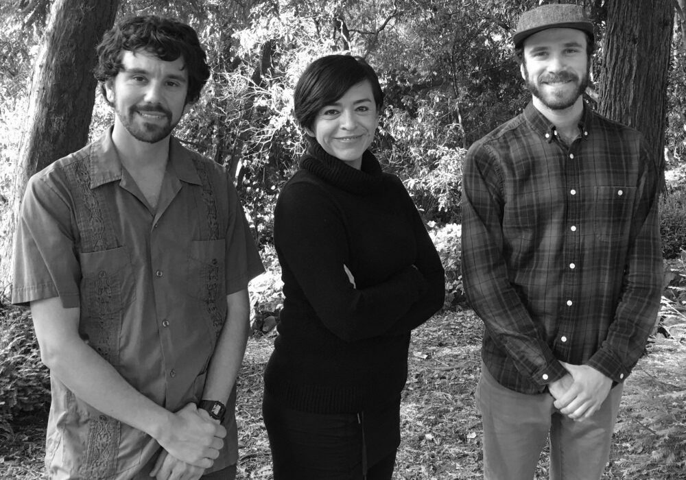 Black and white photo of three people standing outside in front of trees. The person on the left is wearing a short-sleeved shirt, the person in the middle has a black turtleneck with crossed arms, and the person on the right—likely from Berkeley Journalism—is wearing a cap and a plaid shirt.