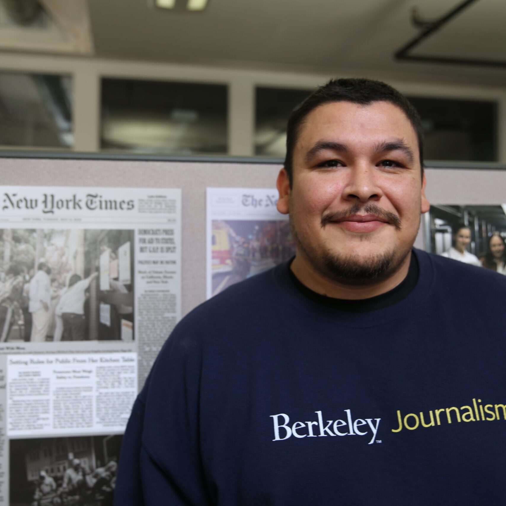 A man in a Berkeley Journalism shirt stands smiling in front of a display featuring The New York Times articles.