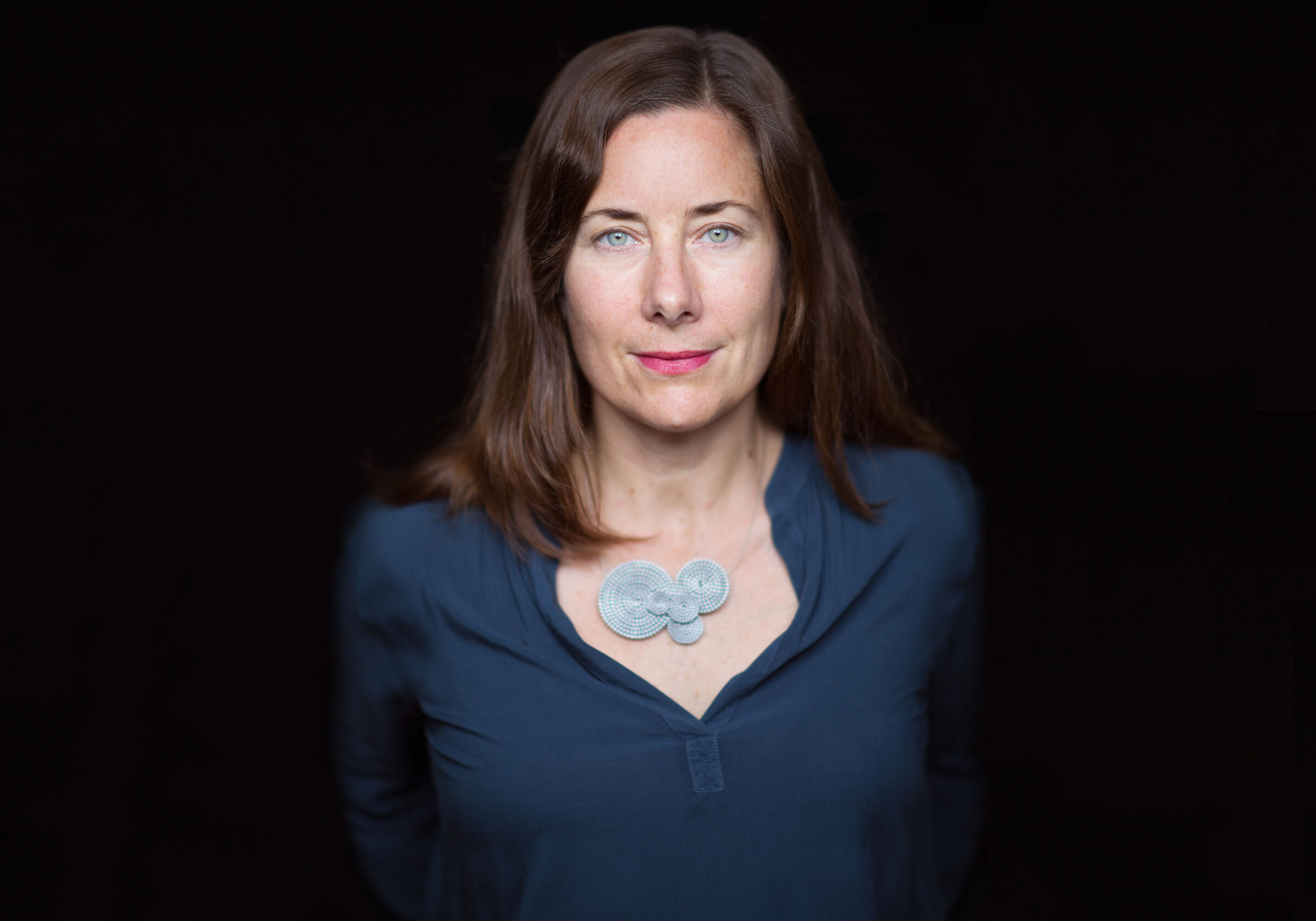 A woman with straight brown hair and blue eyes stands against a dark background. Wearing a navy blue shirt and a large, decorative round necklace in a light color, she looks directly at the camera with a neutral expression that recalls an intensity often seen in Berkeley Journalism alumni.