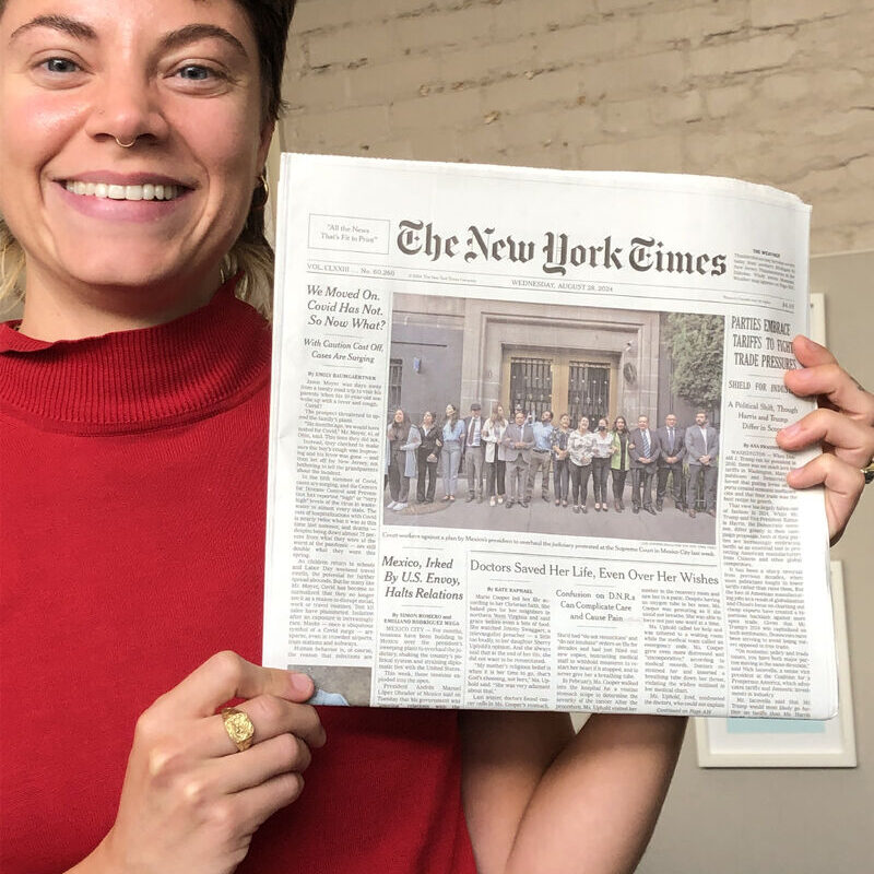 A woman wearing a bright red sweater with short brown hair holds up a print copy of the New York Times with her byline.