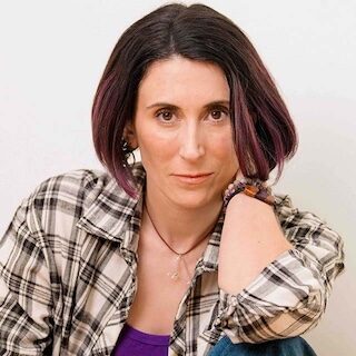 Woman with brown hair in bob cut looking at the camera. She's wearing a plaid shirt opened with a purple shirt underneath,