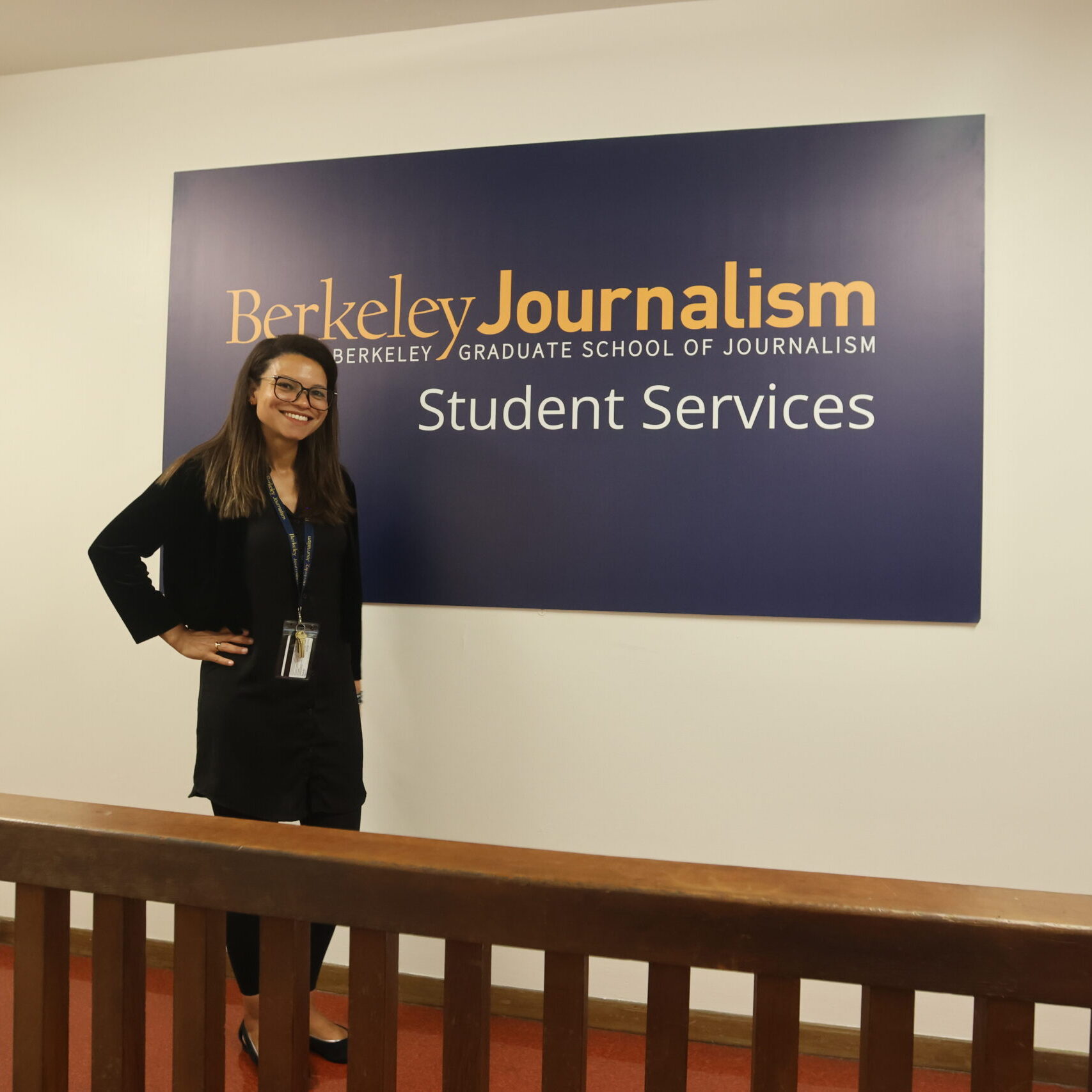 Director of Admissions Director of Journalism dressed all in black with square glasses posed in front of the Berkeley Journalism Student Services sign at the J-School