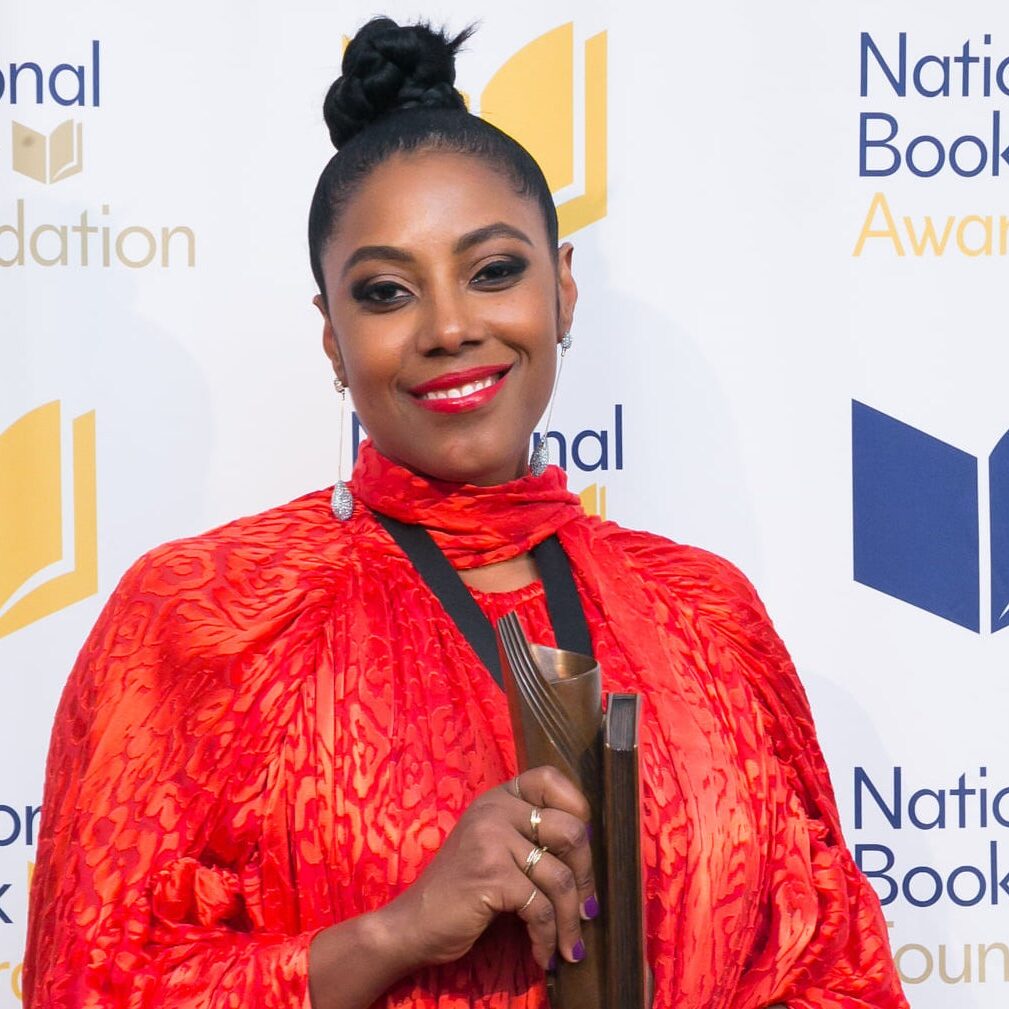 A woman in a vibrant red-orange outfit, a graduate of Berkeley Journalism, holds a National Book Award trophy while smiling at the camera. The backdrop features the logos of the "National Book Foundation" and "National Book Awards".