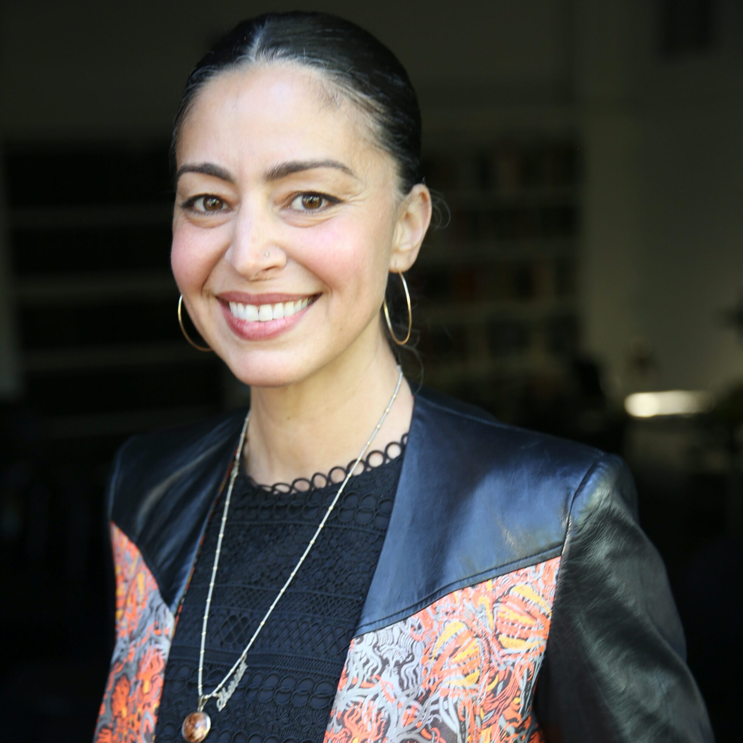 A woman with dark hair pulled back, wearing hoop earrings, a black top with intricate patterns, and a black jacket with colorful textured detail on the shoulders. Shereen Marisol Meraji is smiling and standing indoors against a dark, blurred background.