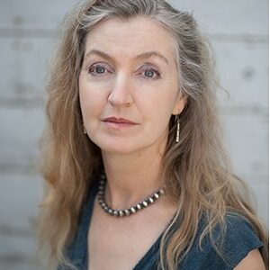 A woman with long, wavy blonde hair and light eyes is wearing a dark V-neck top and a beaded necklace. She has a neutral expression on her face and stands against a light-colored, wooden background, embodying the composed elegance often associated with Berkeley Journalism students.