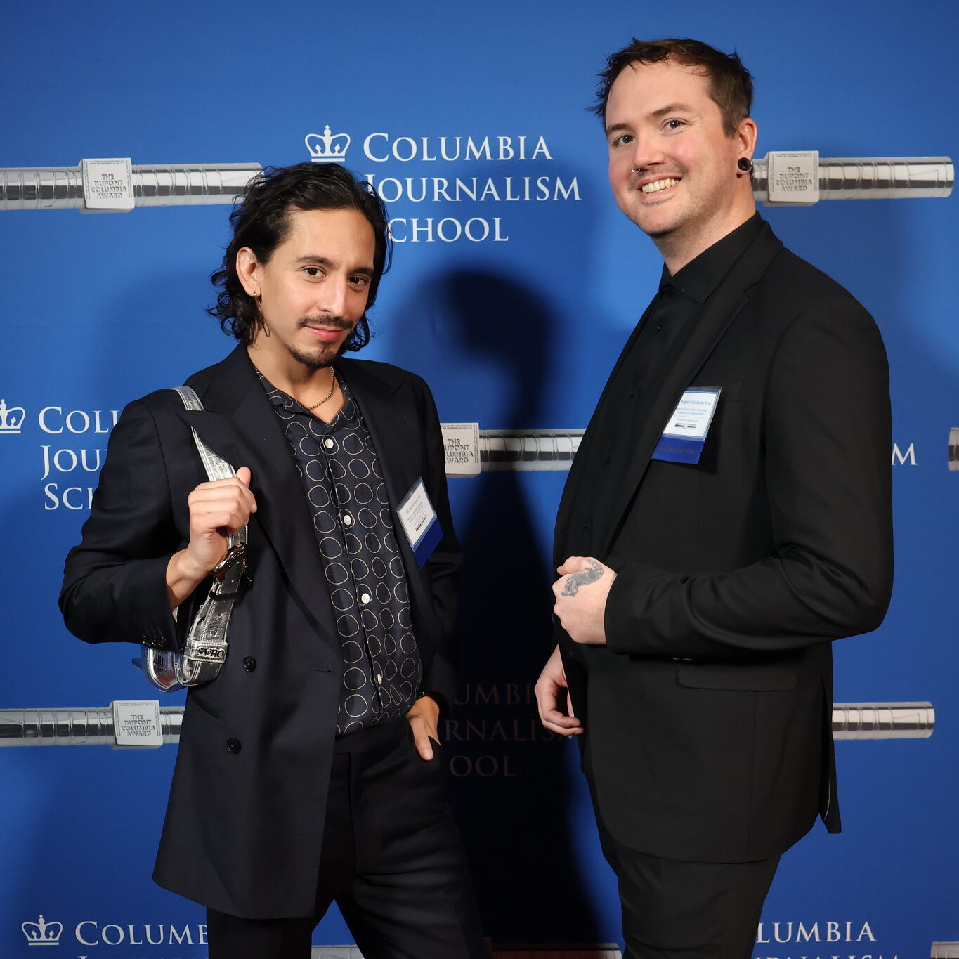 Two mean dressed in black suits on the red carpet smiling with a blue background with white and silver duPont Awards logos.