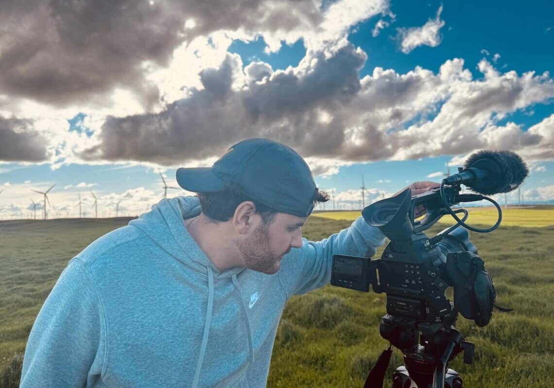 A person in a hoodie and cap operates a video camera in a windy field with wind turbines under a cloudy sky, capturing footage for a climate journalism program.