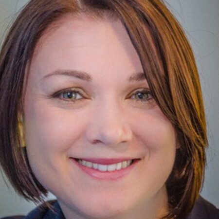 A person with short, brown hair smiles warmly at the camera. The background is blurred with neutral colors, making the person stand out prominently in the image as if they were on a national stage.