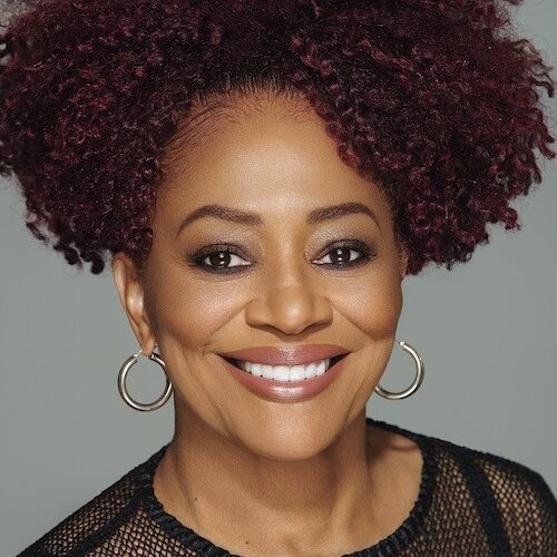 A woman with curly maroon hair, possibly influenced by her time at Berkeley Journalism, smiles warmly. She is wearing a black, semi-transparent, patterned top and large hoop earrings. The background is a solid light gray.