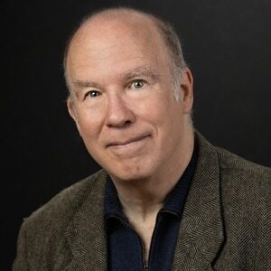 danner Mark Danner, a bald man with light skin and a slight smile, is wearing a gray blazer over a dark zip-up shirt. He is posing against a dark background.